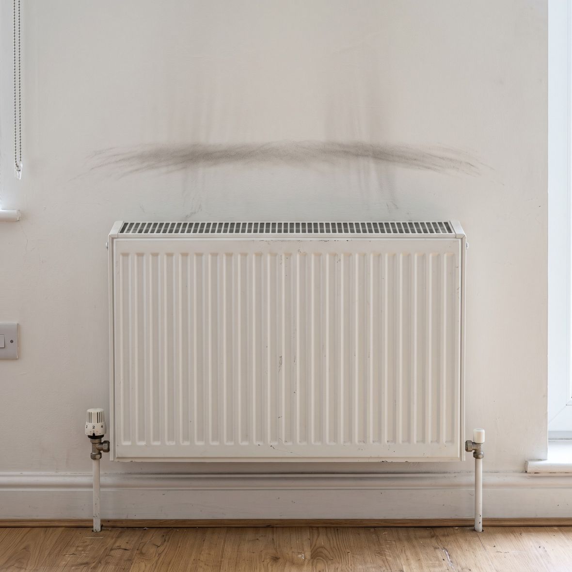 a standard white-painted steel panel heating radiator mounted on a light warm-white painted plaster wall inside a modest, lived-in upper-floor flat in a residential tower block in East London